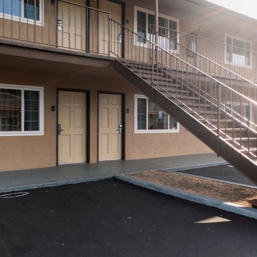 The image shows the exterior of a two-story motel or apartment building with beige walls, several windows, and a set of metal stairs on the right.