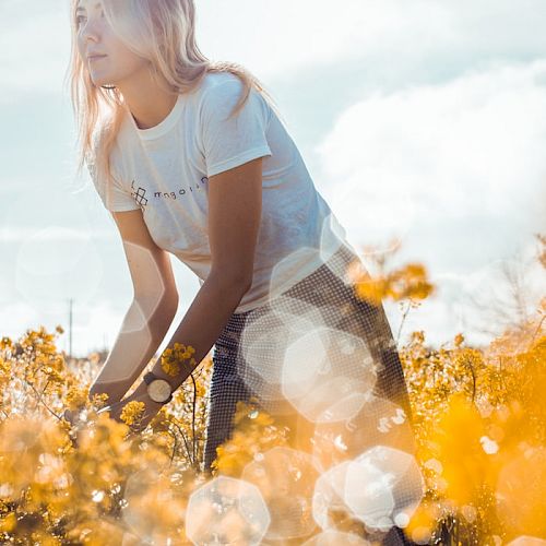 A person is standing in a field of yellow flowers, bending slightly, with sunlight creating a soft, dreamy effect through lens flare.