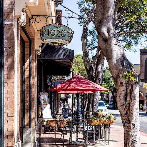 A quaint sidewalk café with red umbrellas, trees lining the street, and a vintage sign above the entrance. Tables and chairs are outside for patrons.