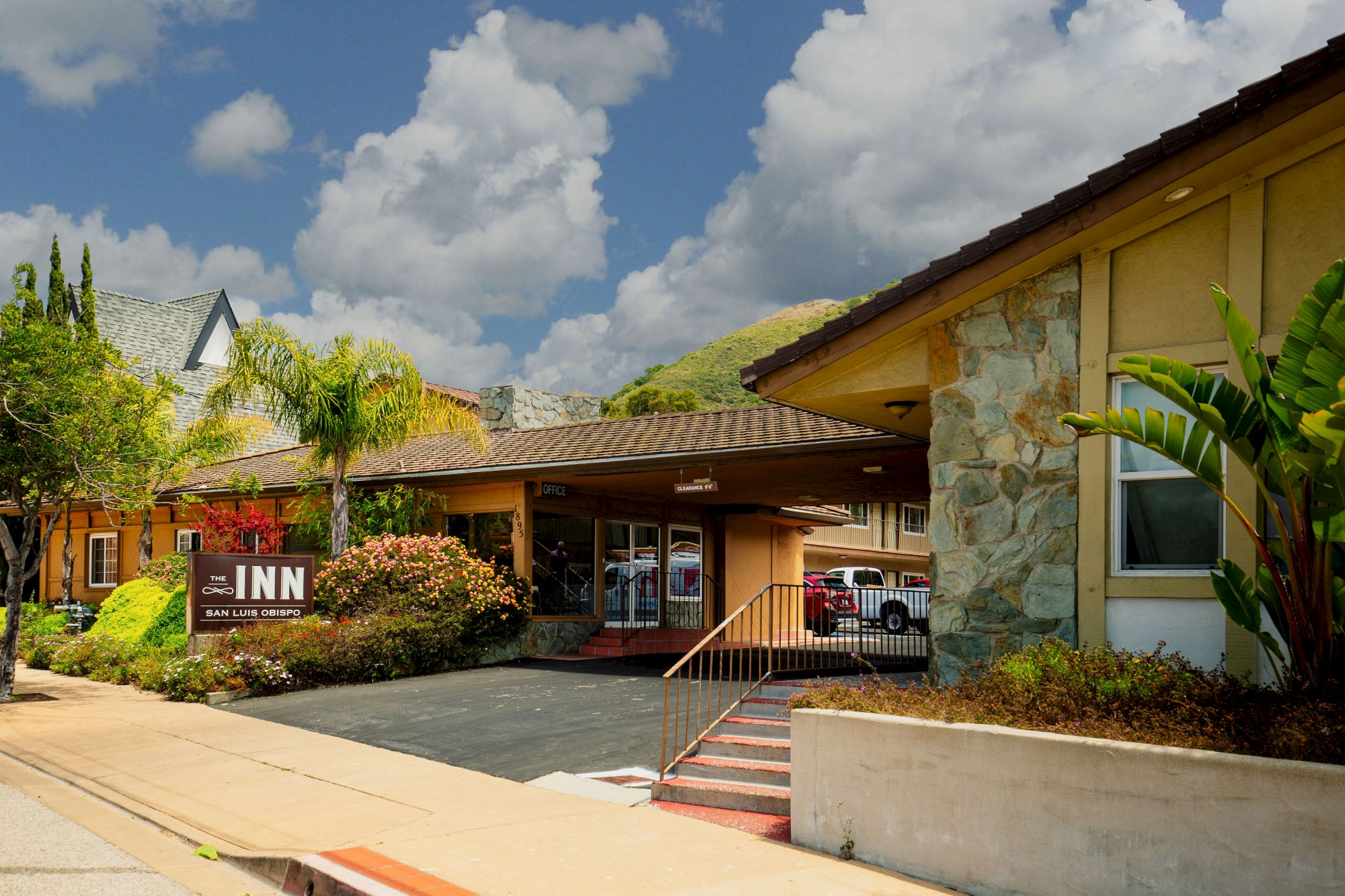 The image shows the facade of an inn with a sign, surrounded by greenery, a stone wall, and a clear sky with clouds.