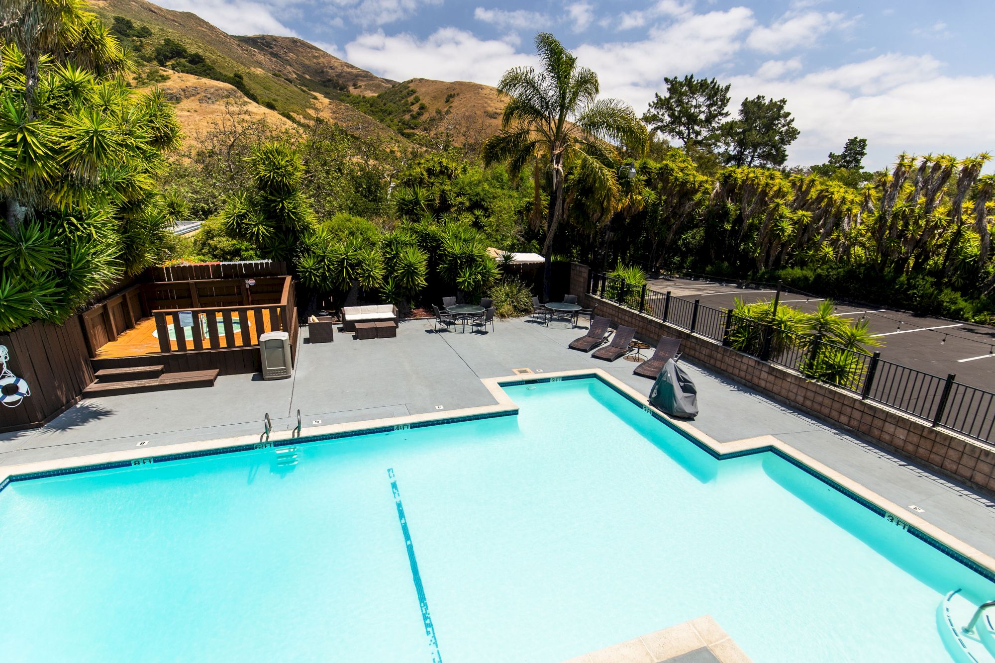 The image shows a spacious outdoor pool area with lounge chairs, surrounded by lush greenery and hills in the background under a partly cloudy sky.