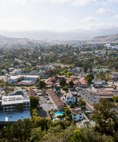 A sunny hillside cityscape with a mix of houses, trees, and low-rise buildings stretching toward rolling hills in the distance, under a bright sky.