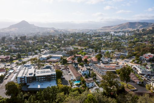 A sunny hillside cityscape with a mix of houses, trees, and low-rise buildings stretching toward rolling hills in the distance, under a bright sky.