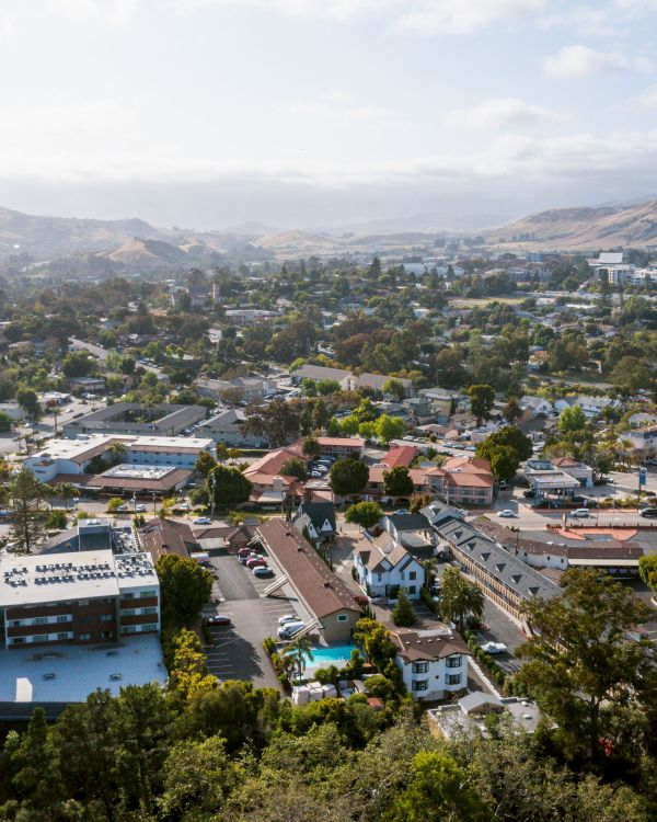 Aerial view of a suburban area with various buildings, greenery, and a mountain in the background under a partly cloudy sky.