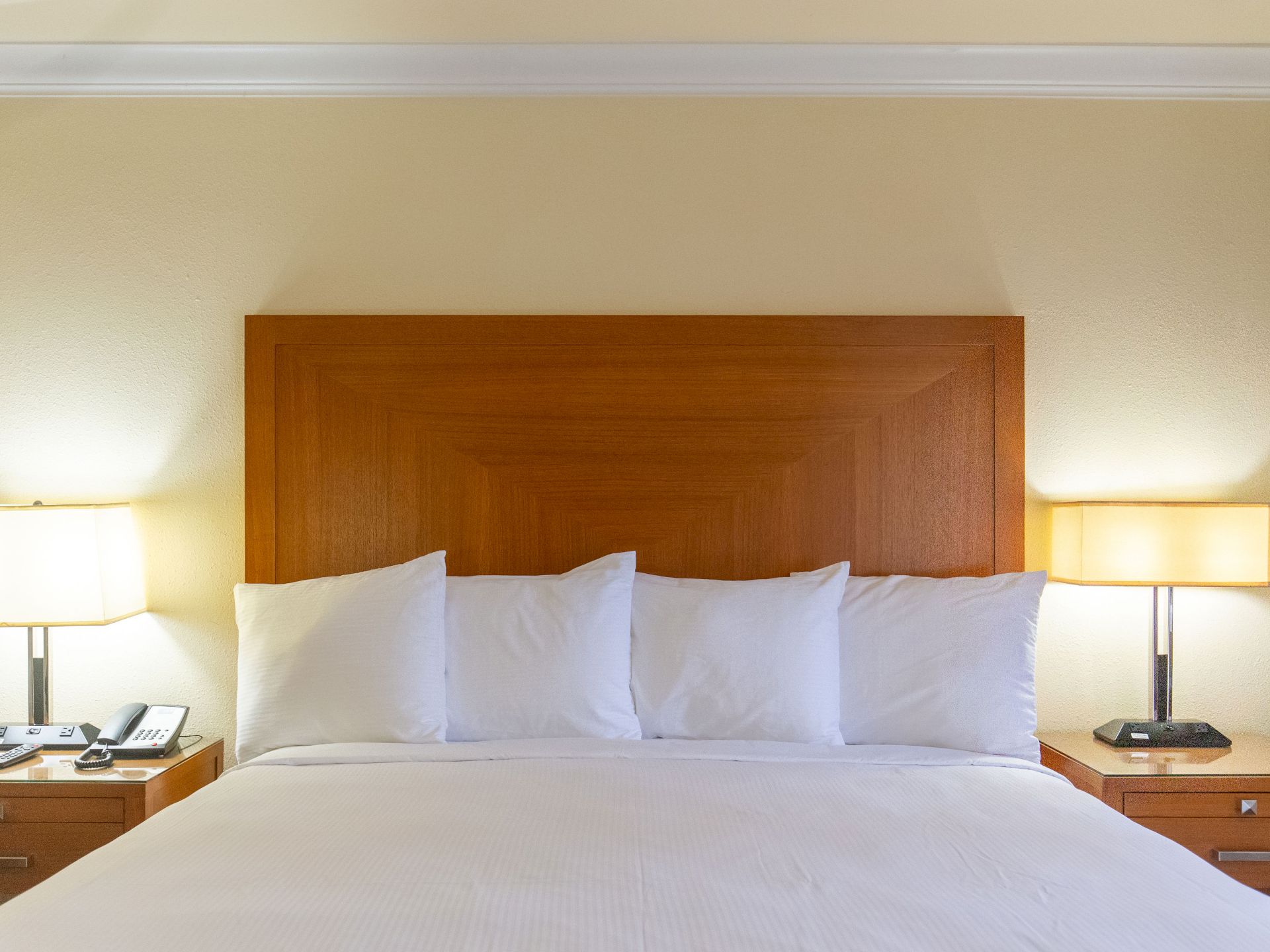 A hotel room bed with a wooden headboard, white bedding, and pillows, flanked by nightstands with lamps.
