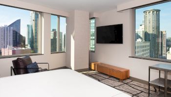 The image shows a modern hotel room with a large bed, a wall-mounted TV, floor-to-ceiling windows, and a view of city skyscrapers, ending the sentence.