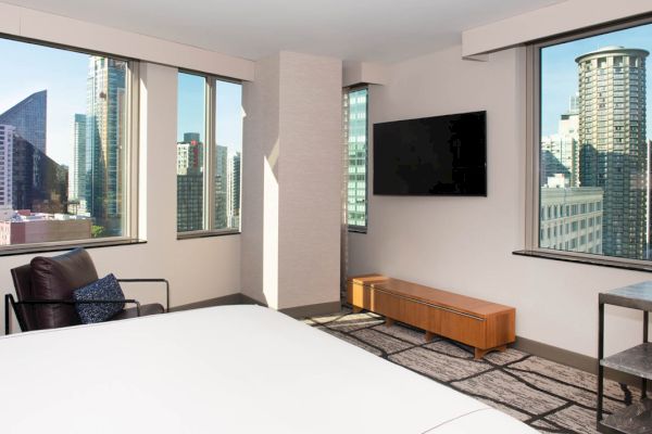 A hotel room with a city view, featuring a bed, chair, TV, and windows.