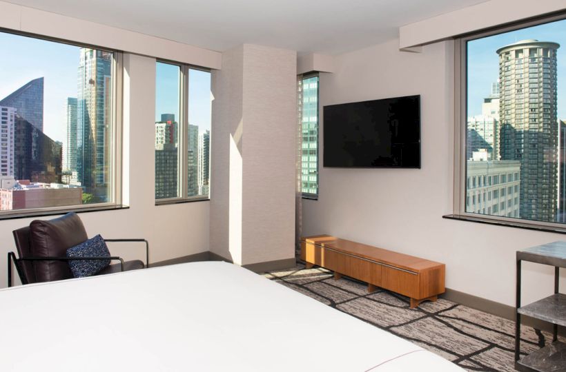 The image shows a modern hotel room with a large bed, a wall-mounted TV, floor-to-ceiling windows, and a view of city skyscrapers, ending the sentence.