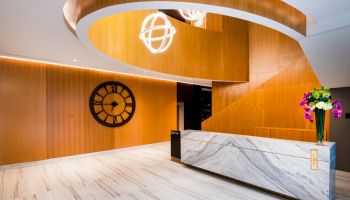 This image shows a modern lobby with a marble reception desk, a large wall clock, stylish lighting fixtures, and a bouquet of flowers on the desk.