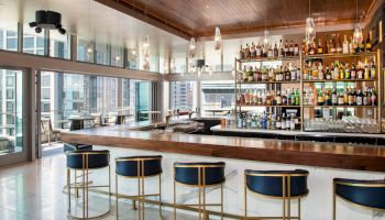 This image shows a modern bar with high chairs, a wide counter, and a well-stocked shelf of liquor bottles beneath hanging lights.