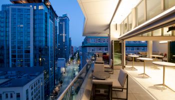 The image shows a modern cityscape at dusk with tall buildings. A balcony with tables and chairs overlooks a street lined with lights.
