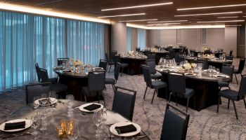 The image shows a banquet hall setup with round tables adorned with black napkins, glassware, and floral centerpieces, under modern lighting.