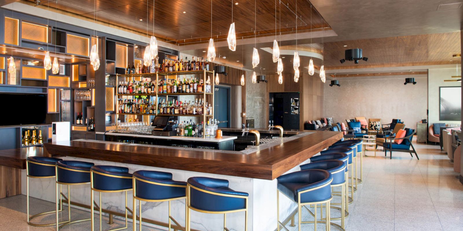 The image shows a modern bar with a long counter, blue bar stools, hanging lights, and a well-stocked display of bottles behind the counter.