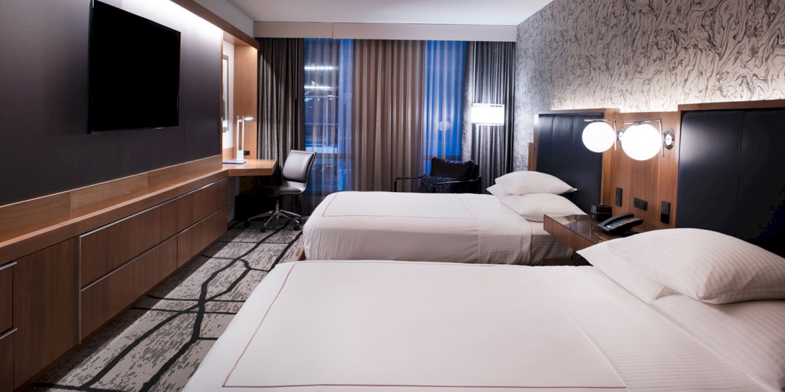 A modern hotel room with two neatly made beds, a wall-mounted TV, a desk with a chair, and stylish decor, including a patterned wall and carpet.