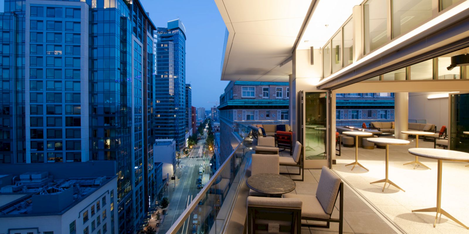 An outdoor terrace overlooking a cityscape with modern buildings under a twilight sky. The terrace has chairs and tables arranged neatly.