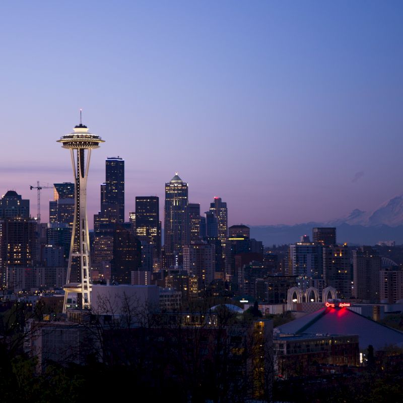 A view of a city skyline at dusk, with the iconic Space Needle prominently featured, mountains in the background, and buildings lit up.
