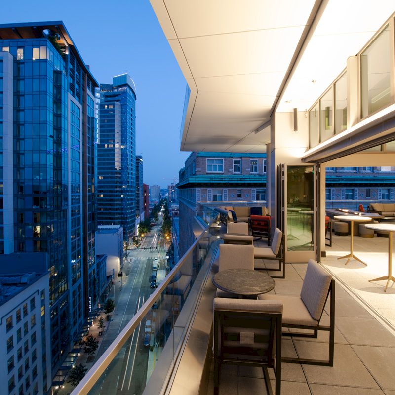 A modern balcony overlooks an urban cityscape during dusk, featuring tables, chairs, and a well-lit interior space with large windows.