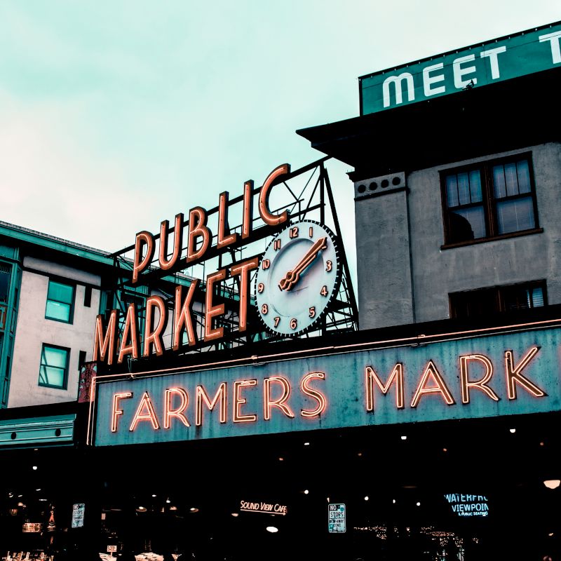The image shows the iconic neon sign of a public market and a farmer's market, with buildings and a partially visible sign saying "MEET THE."