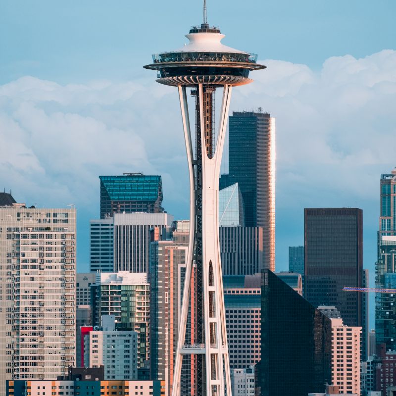 The image shows the Space Needle towering above the Seattle skyline, surrounded by various modern buildings and skyscrapers under a partly cloudy sky.