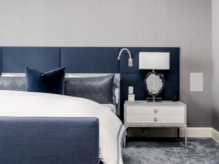 A modern bedroom features a bed with blue headboard and pillows, a nightstand with a lamp, a wall-mounted light, and a decorative object.
