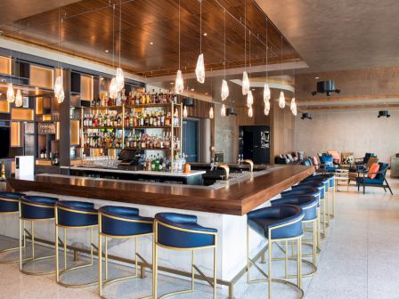 The image shows a modern bar area with a wooden counter, blue chairs, hanging lights, and a well-stocked bar, with a seating area in the background.