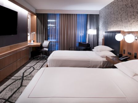 The image shows a modern hotel room with two beds, a large flat-screen TV, a desk with a chair, and stylish decor.