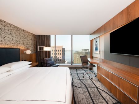 A modern hotel room features a large bed, wall-mounted TV, desk with a chair, and a view of city buildings through floor-to-ceiling windows.