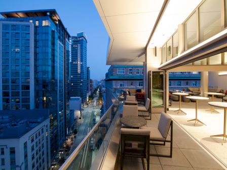 The image shows a modern cityscape viewed from a high balcony with tables and chairs, overlooking tall buildings and a street below at dusk.