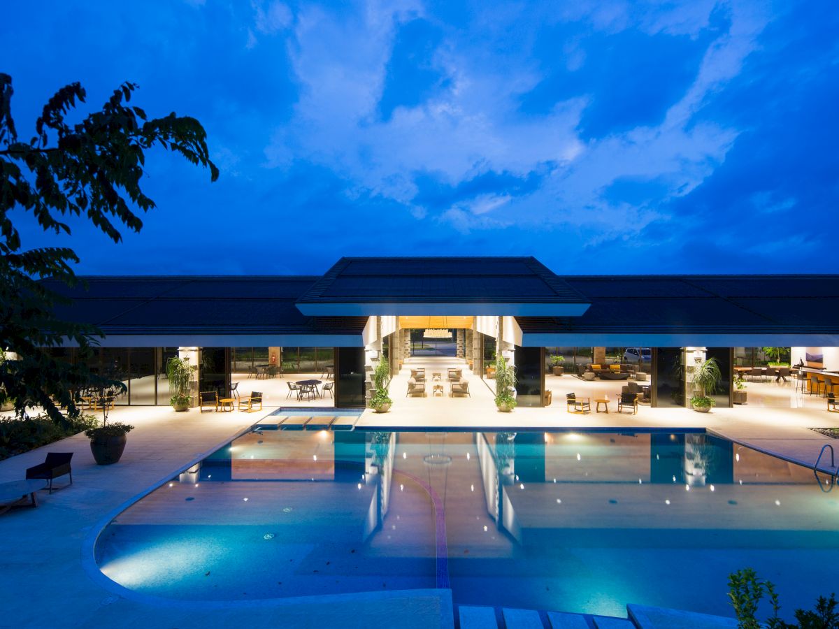 An illuminated luxury resort with a large swimming pool reflecting the lights, surrounded by lounge areas and open interior spaces, under a dusky sky.