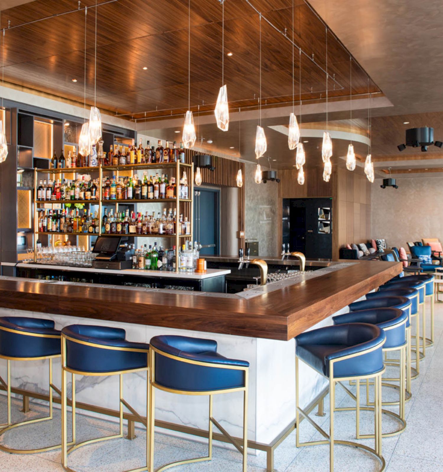 A modern bar with a central counter, numerous stools, hanging lights, and a well-stocked liquor shelf. The space is spacious and stylish.
