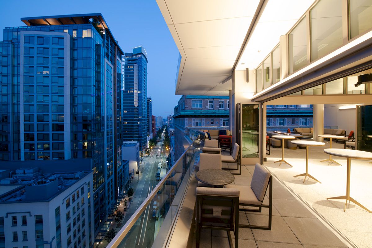 A modern cityscape with tall buildings at dusk, featuring a spacious balcony with outdoor seating and tables.