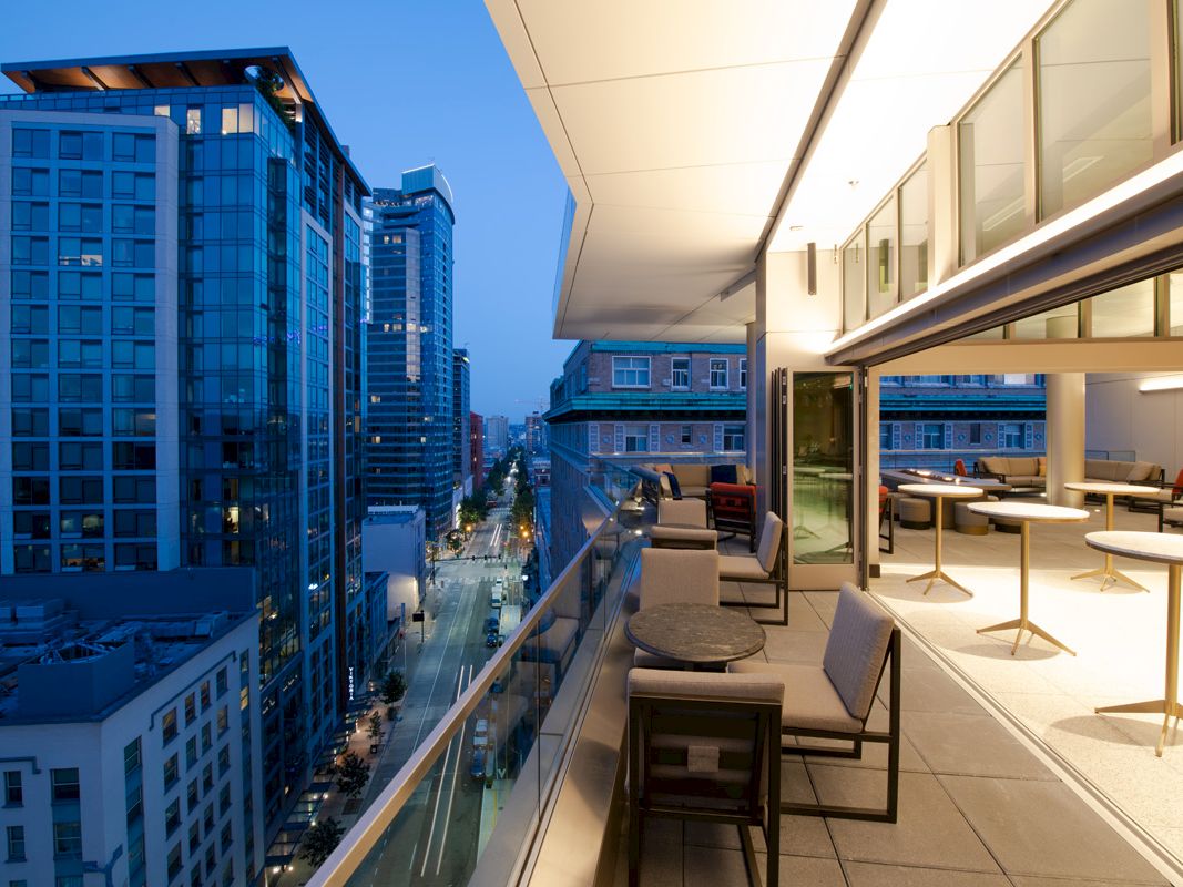 A modern cityscape with tall buildings at dusk, featuring a spacious balcony with outdoor seating and tables.