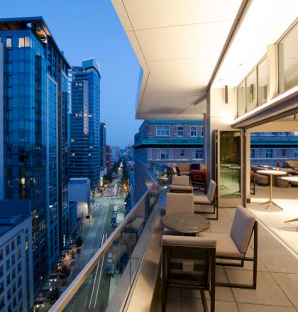 A modern balcony overlooking a cityscape at dusk, with seating areas and tables, adjacent to a stylish indoor space with large windows.