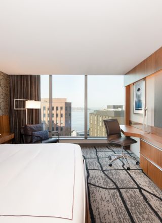 A modern hotel room features a large bed, a wall-mounted TV, a desk with a chair, and a window with a view of buildings and the water outside.