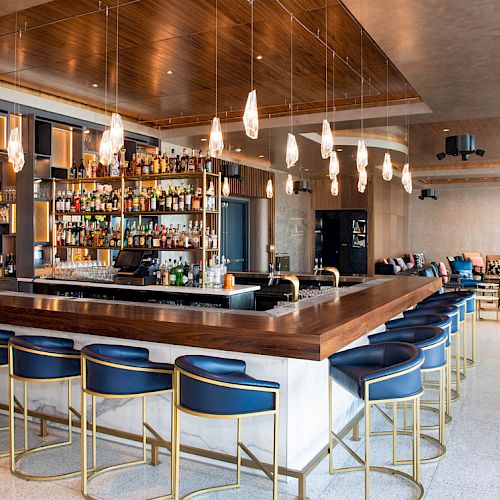 A modern bar with a wooden countertop, blue and gold bar stools, a well-stocked back bar, and contemporary pendant lighting.