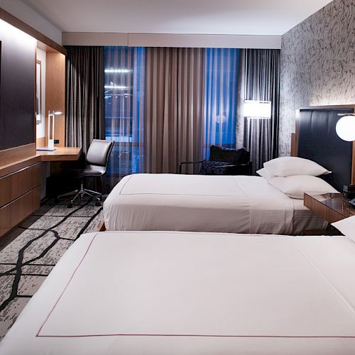 The image shows a modern hotel room with two neatly made beds, a wall-mounted TV, a desk with a chair, and stylish decor.