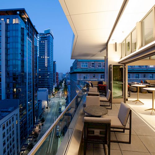 A balcony with seating overlooks a cityscape with tall buildings, illuminated at dusk. Tables and chairs are set up inside and outside.