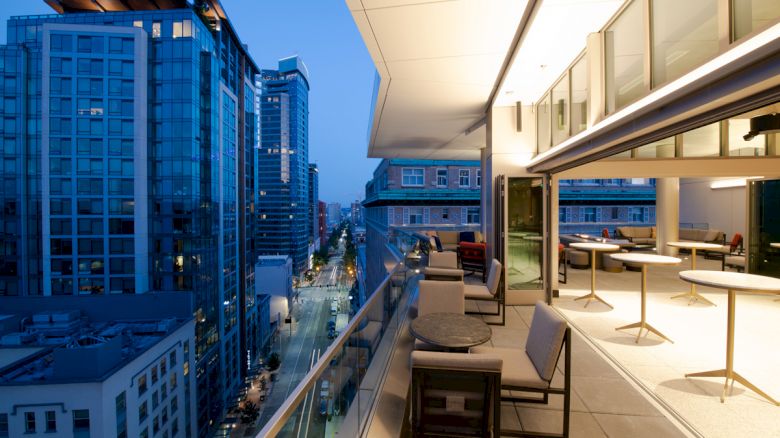 A modern outdoor balcony with tables and chairs overlooks a city street lined with high-rise buildings during twilight, featuring city lights.