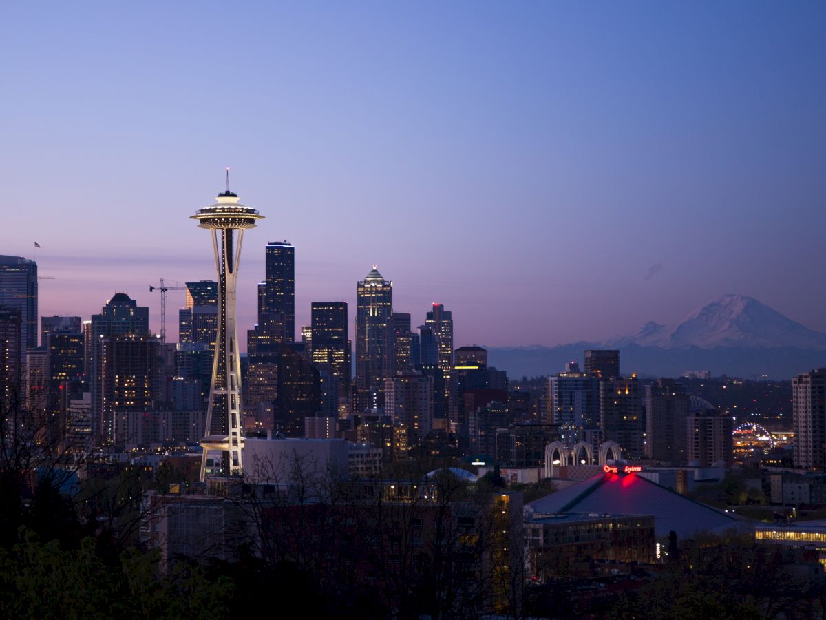 The image shows a twilight view of a city's skyline, featuring a prominent tower and a mountain in the background. The city is Seattle, Washington.