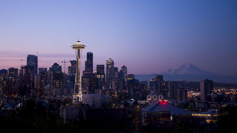 The image shows a twilight view of a city's skyline, featuring a prominent tower and a mountain in the background. The city is Seattle, Washington.