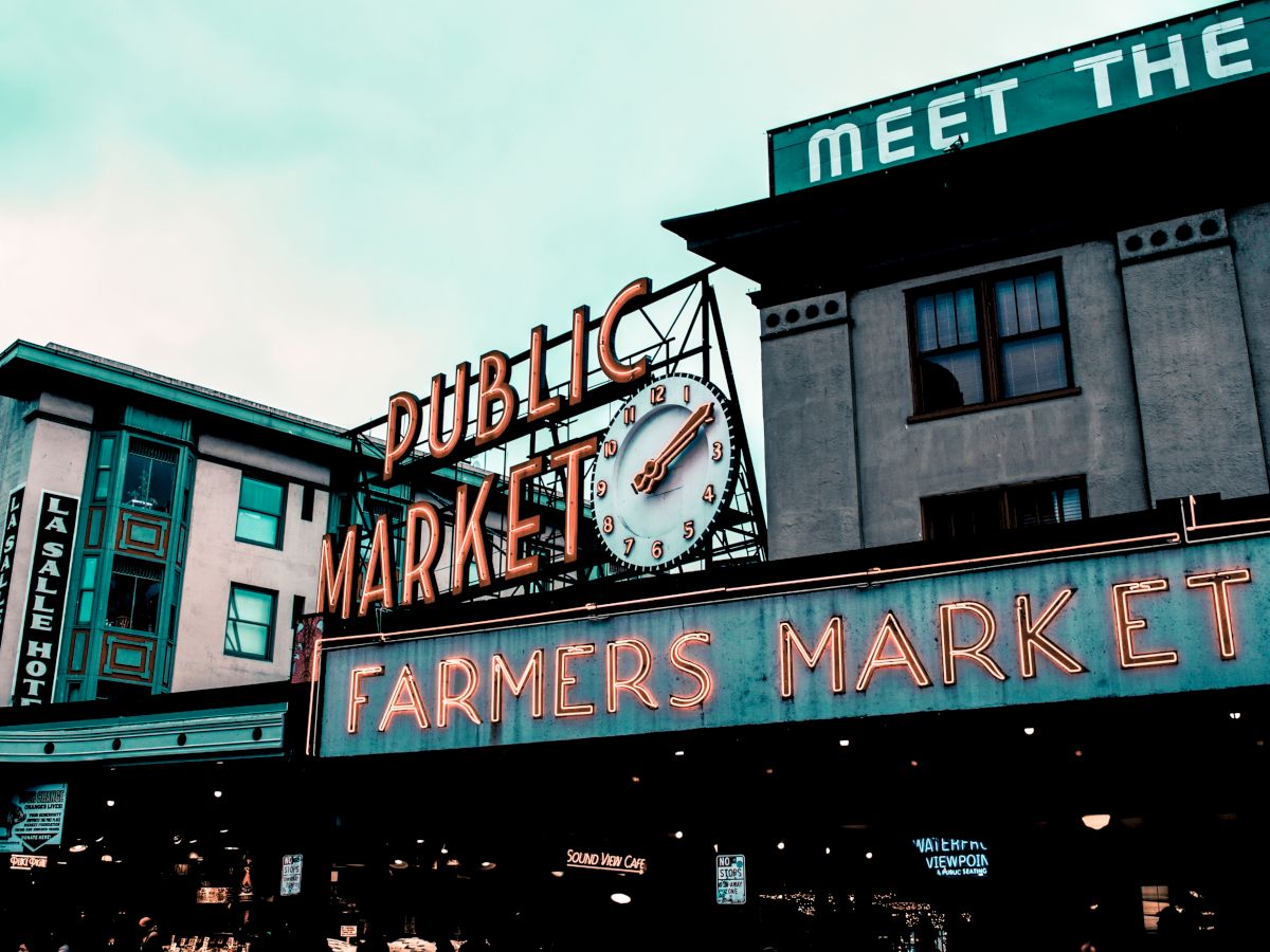 The image shows the iconic neon signage for a Public Market and a Farmers Market against the backdrop of urban buildings under a cloudy sky.
