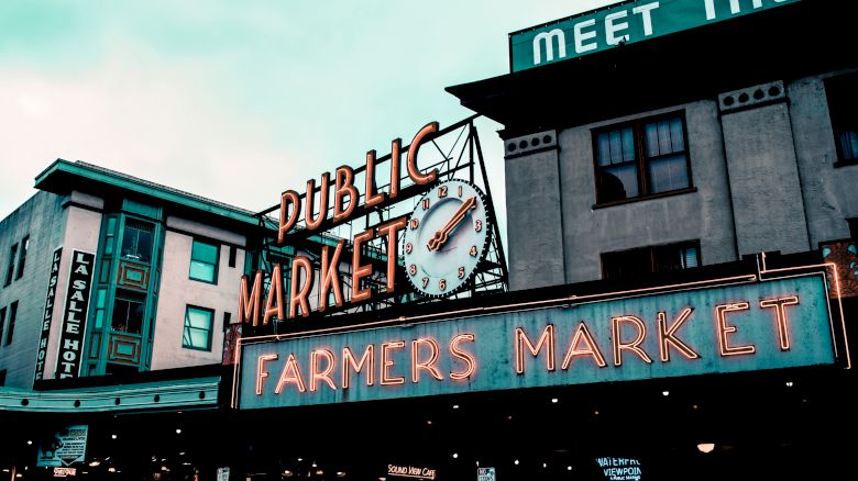 The image shows the iconic neon signage for a Public Market and a Farmers Market against the backdrop of urban buildings under a cloudy sky.