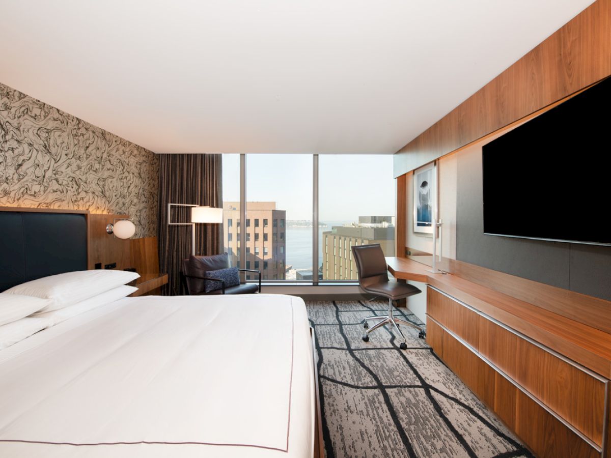 This image shows a modern hotel room with a large bed, wall-mounted TV, desk, chair, floor-to-ceiling windows, and a view of city buildings.