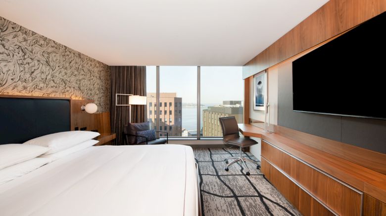 This image shows a modern hotel room with a large bed, wall-mounted TV, desk, chair, floor-to-ceiling windows, and a view of city buildings.