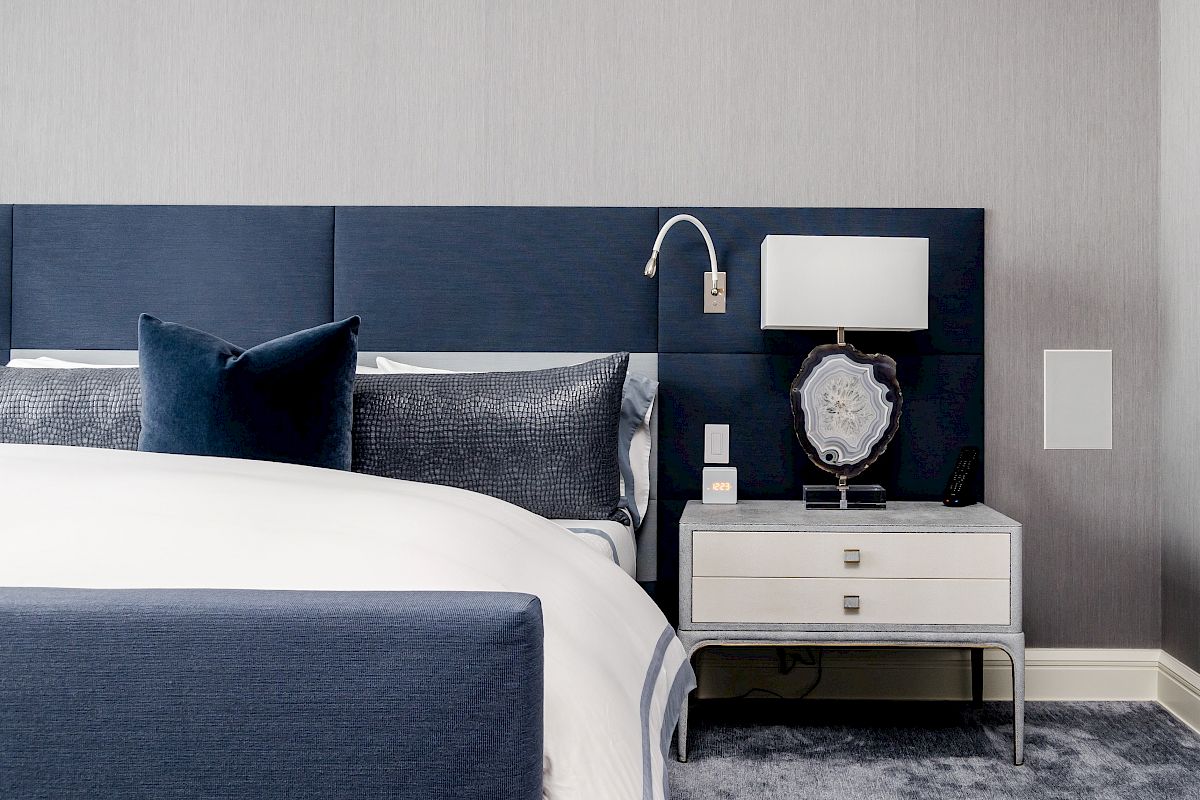 A modern bedroom features a blue upholstered bed, nightstand with a lamp and decor, gray wall, and carpeted floor.
