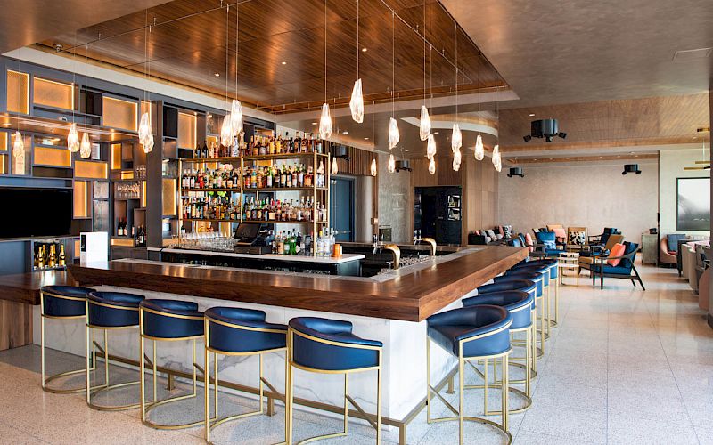 The image shows a modern bar with a wooden counter, blue bar stools, shelves stocked with bottles, hanging lights, and a spacious seating area.