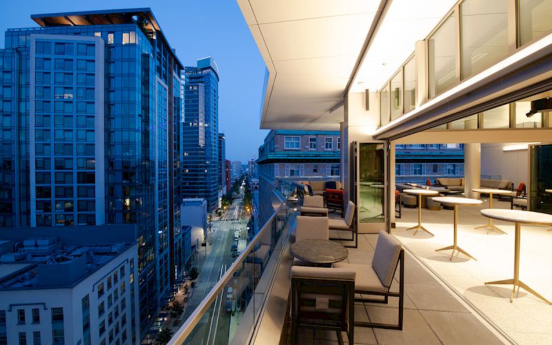 A modern balcony with seating overlooks an urban street with tall buildings at dusk. Lights illuminate the balcony and interior space.