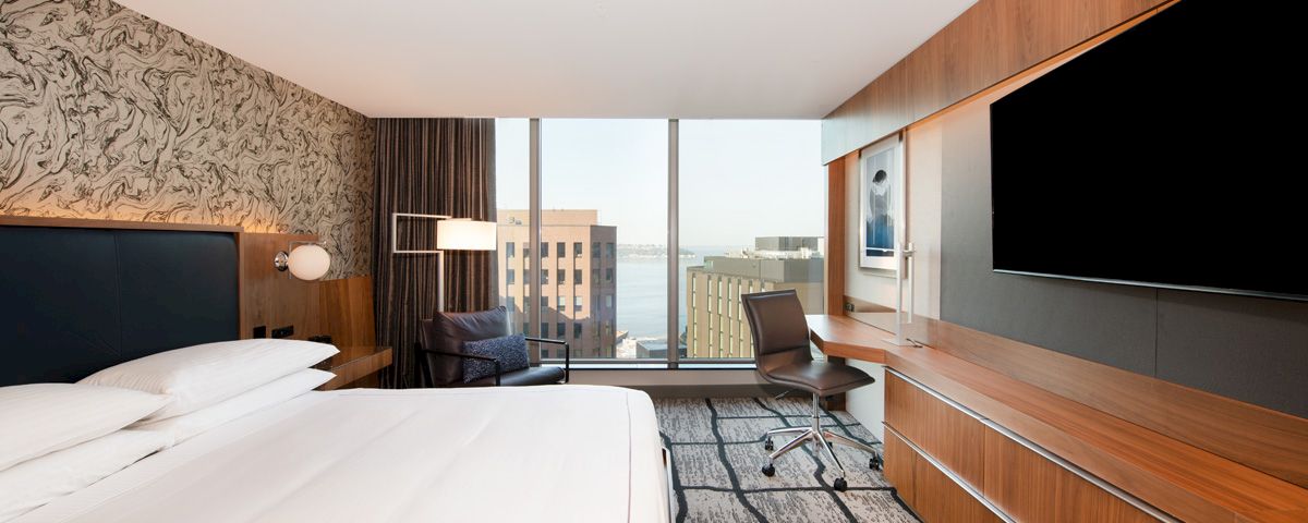 A hotel room with a bed, desk, chair, and TV, overlooking the city.