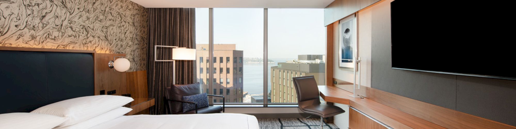 A modern hotel room features a king-sized bed, a large flat-screen TV, a work desk, an armchair, and a window with a city view.