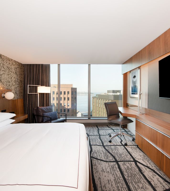 A hotel room with a bed, desk, chair, and TV, and a view of the city through a large window.